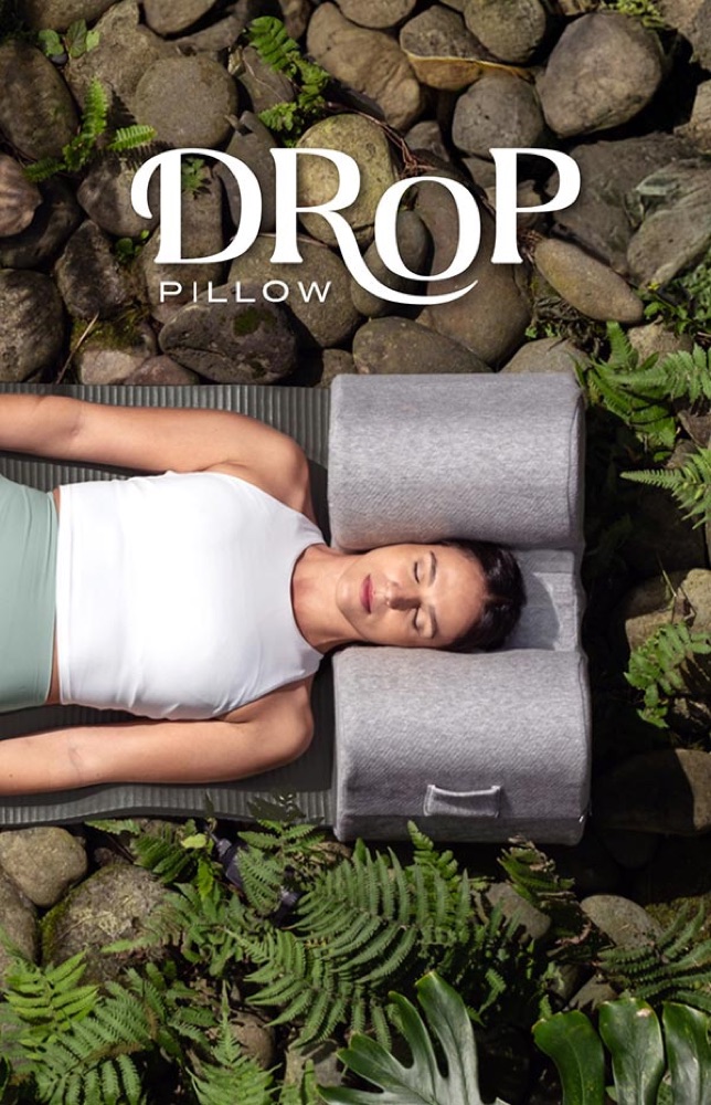 Seen from above: a woman lying on a mat among ferns and river stones, her head resting between the two arms of the Drop Pillow