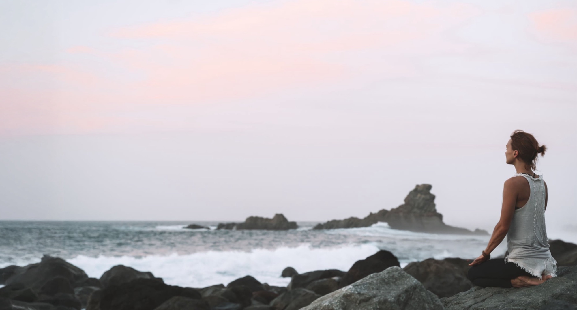 A person sitting cross-legged on rocks above the sea at first light